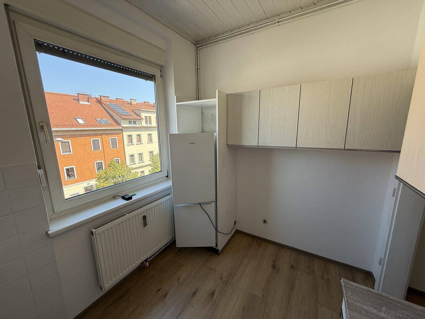 Helle Küche mit Fenster, Kühlschrank und Hängeschränken, bietet einen Blick auf die umliegenden Dächer.