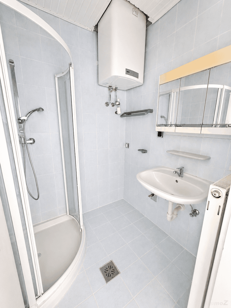 Badezimmer mit Duschkabine, Waschbecken, Spiegelschrank und hellblauen Fliesen, inklusive Warmwasserboiler.