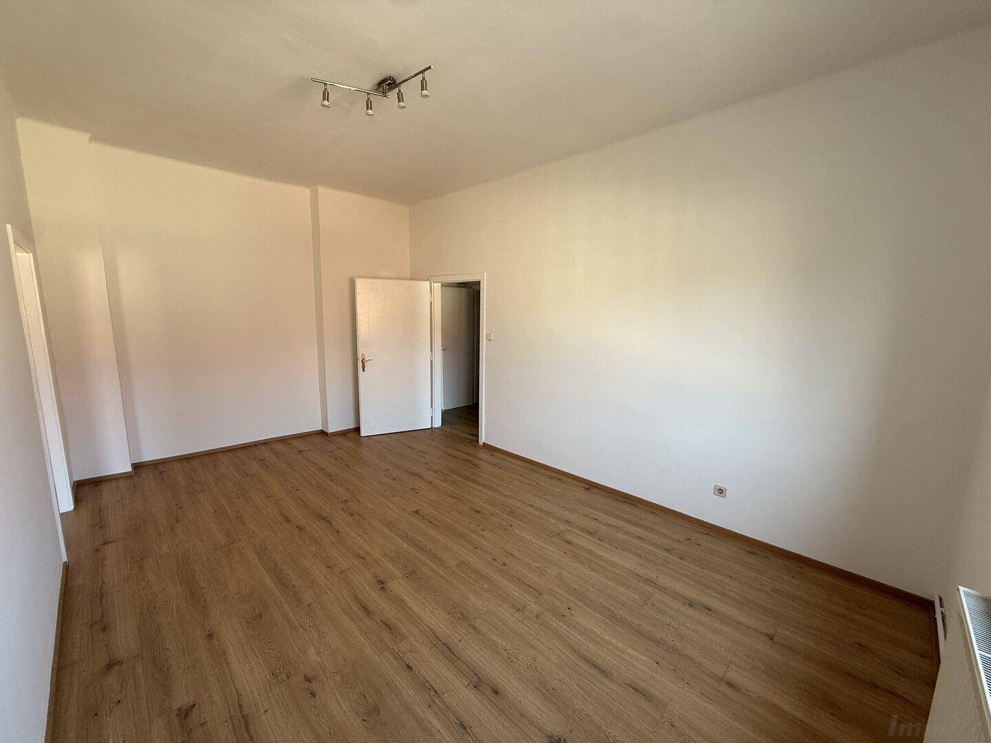 Geräumiges, leeres Zimmer mit hellem Laminatboden und weiß gestrichenen Wänden, vielseitig nutzbar.