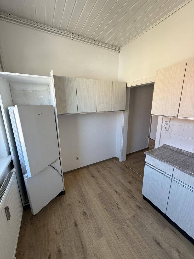 Küchenbereich mit Kühlschrank und Hängeschränken, bietet Stauraum und Zugang zu weiteren Räumen.