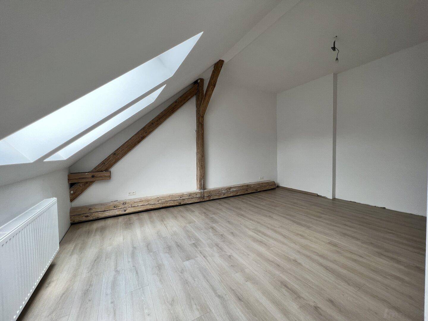 Dachgeschossraum mit sichtbaren Holzbalken, schrägen Wänden und zwei Dachfenstern.