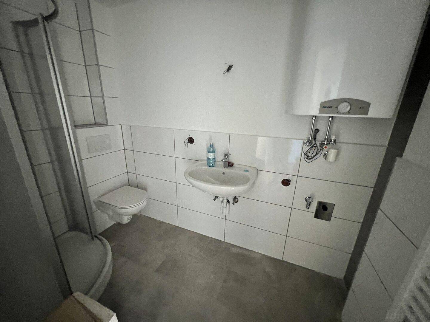Funktionales Badezimmer mit weißen Fliesen, Dusche, WC und Warmwasserboiler.