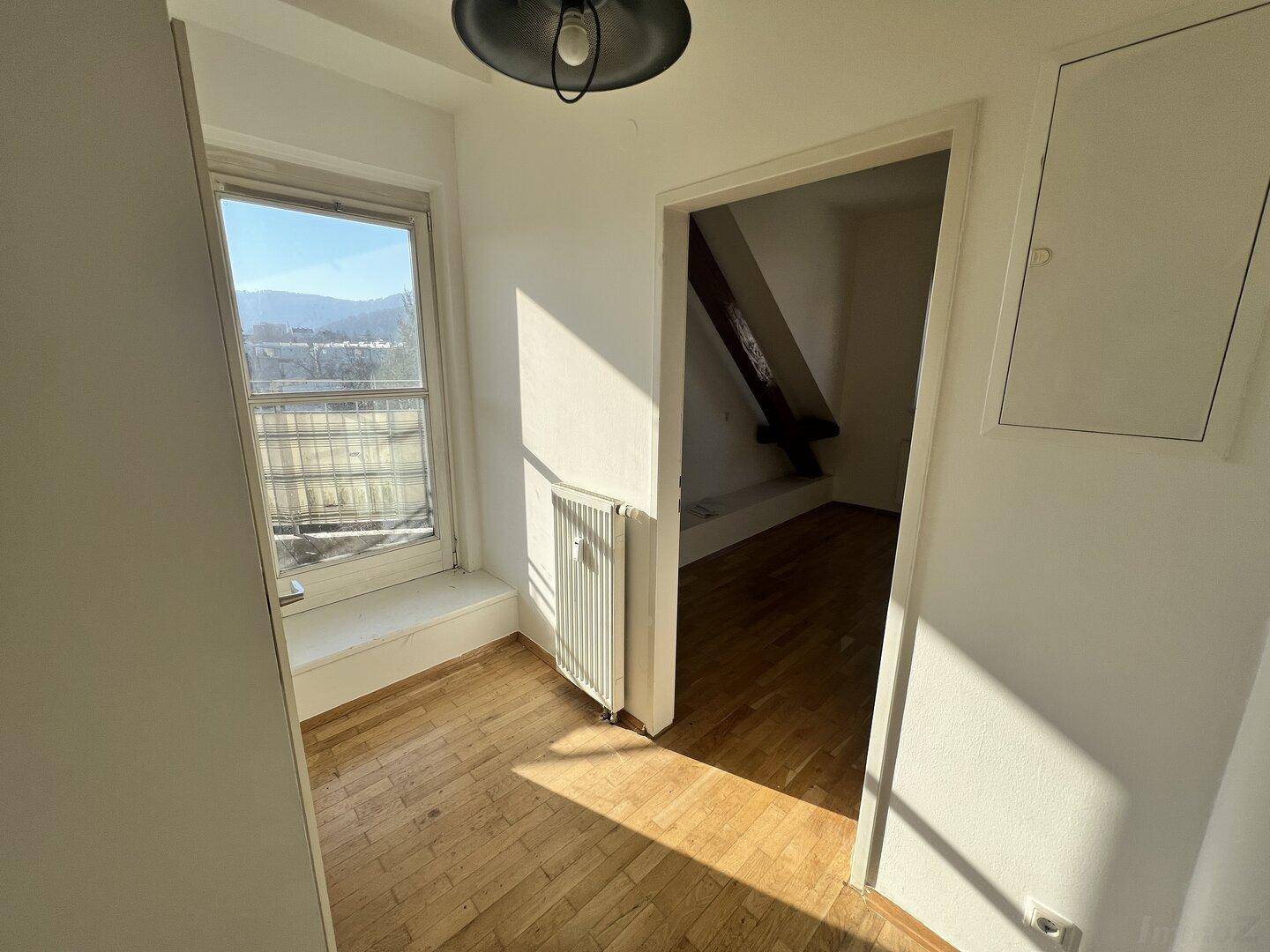 Heller Flur mit Parkettboden, Fenster und Zugang zu einem weiteren Zimmer im Dachgeschoss.