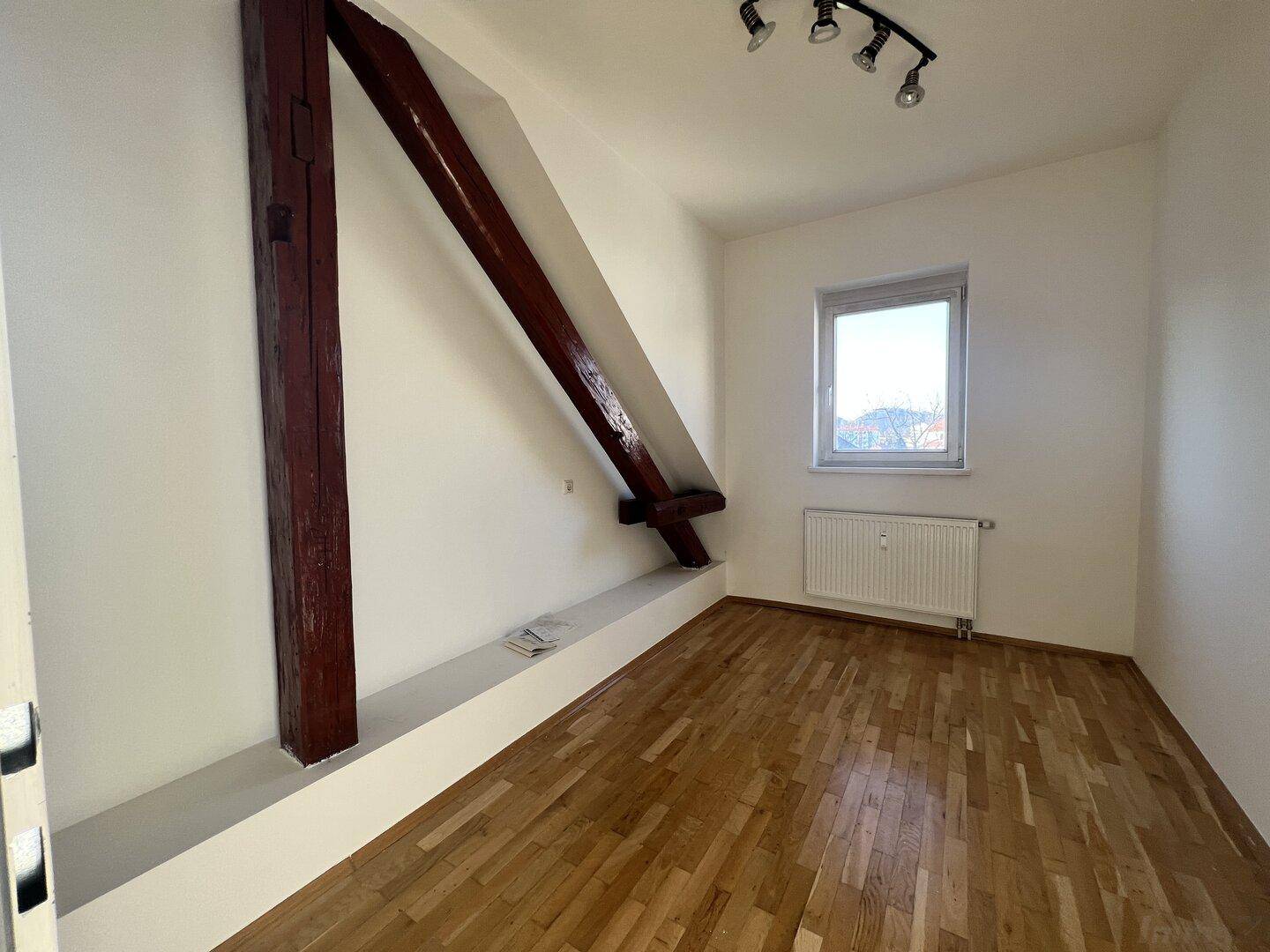 Geräumiges Dachzimmer mit sichtbaren Holzbalken, Parkettboden und einem Fenster für Tageslicht.