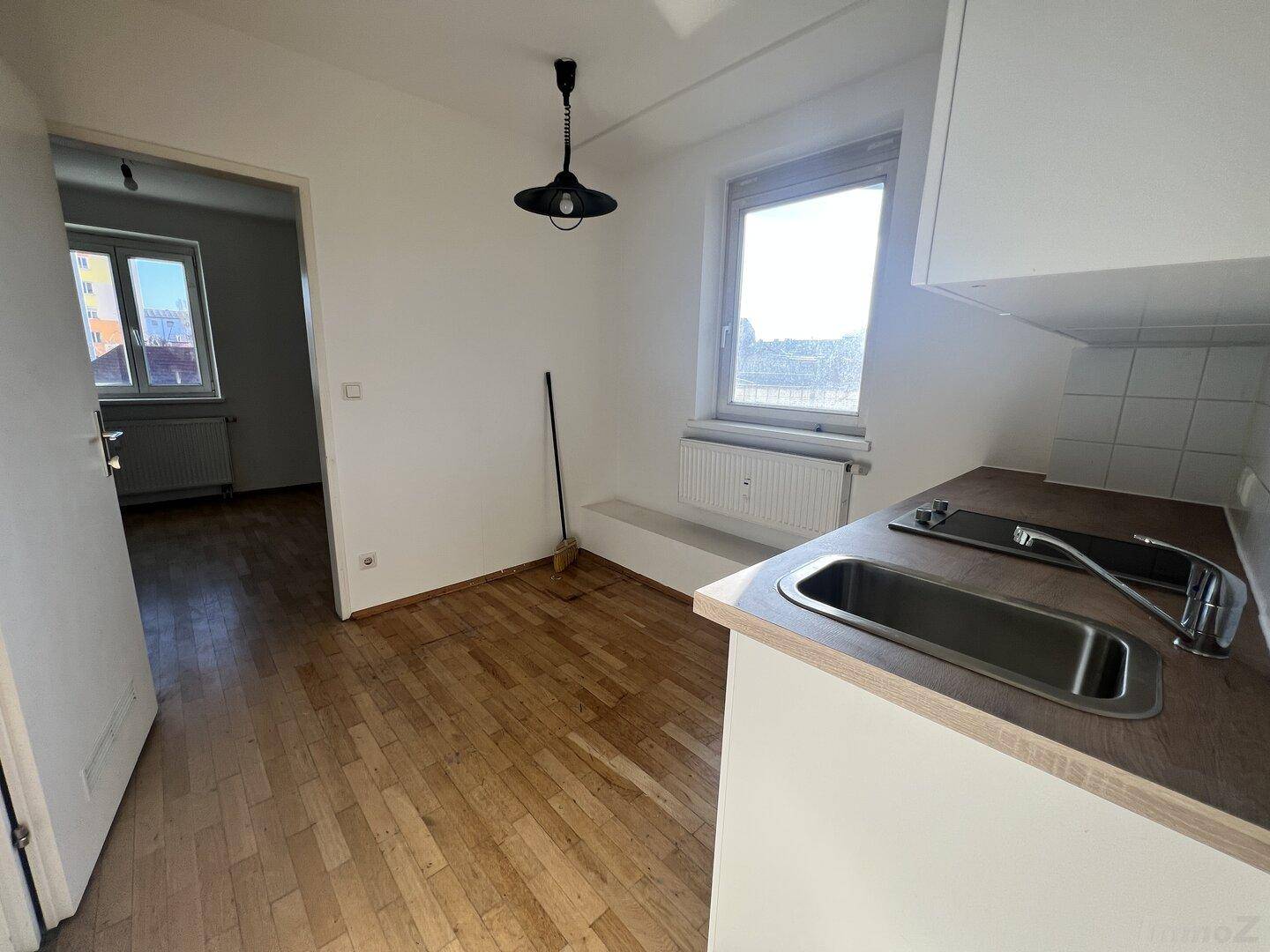 Heller Raum mit Parkettboden und einer kleinen Küchenzeile, offen zu einem weiteren Zimmer.