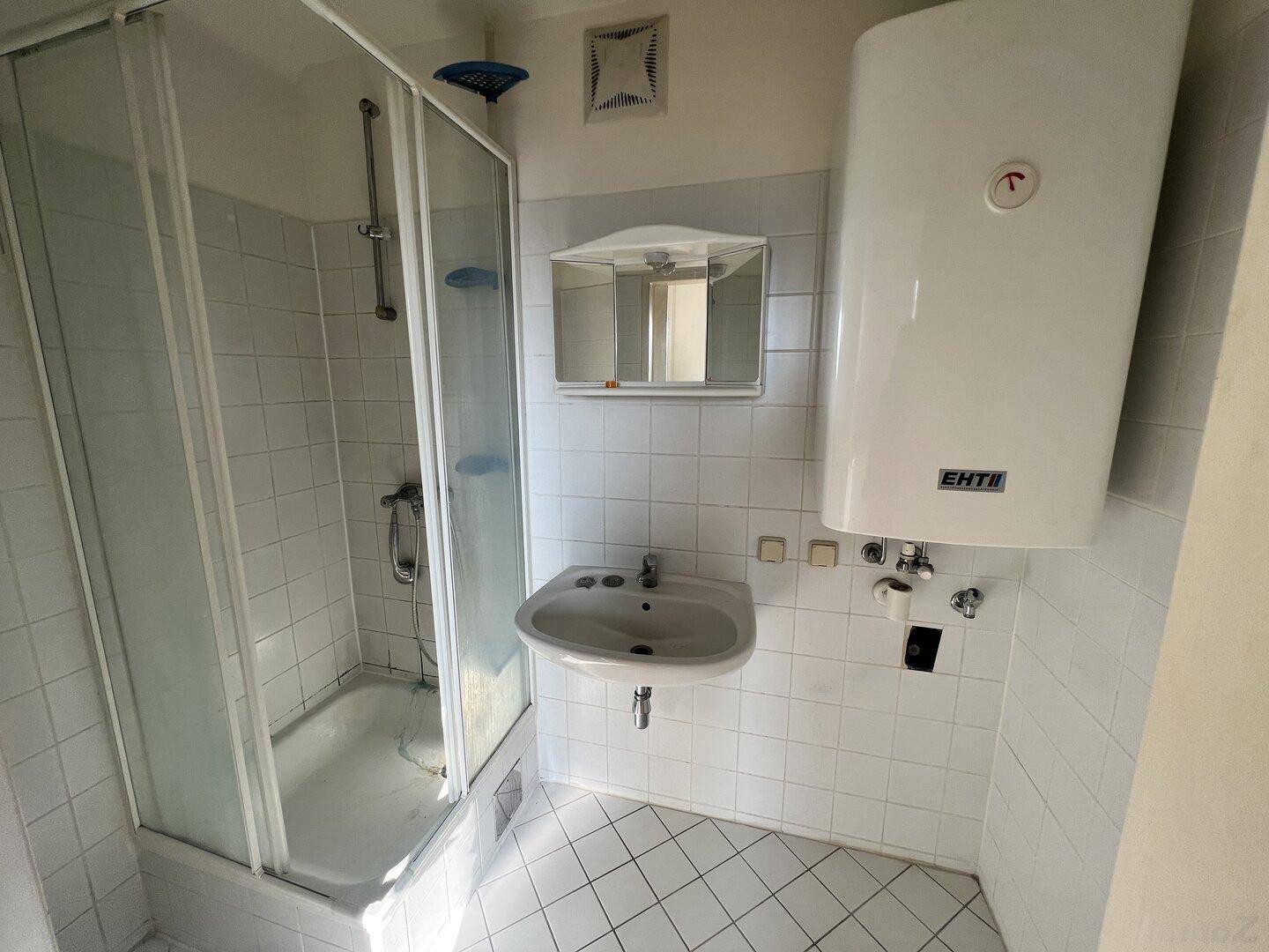 Helles Badezimmer mit weißen Fliesen, Duschkabine, Waschbecken und Warmwasserboiler.