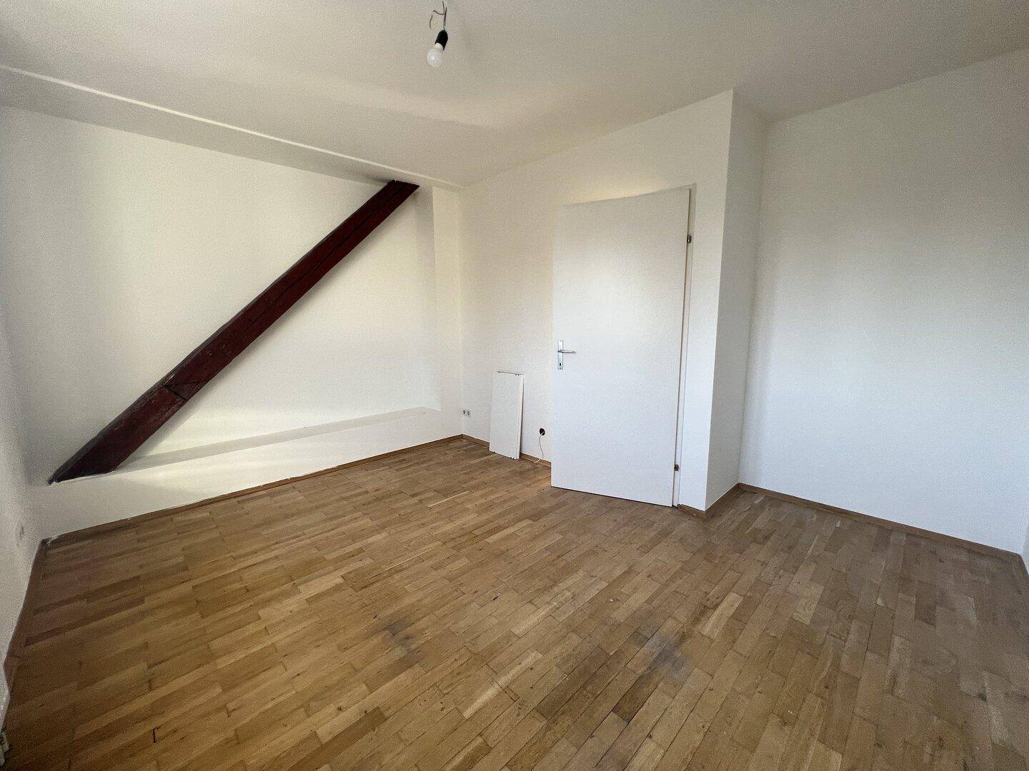 Geräumiges Zimmer im Dachgeschoss mit Parkettboden, weißen Wänden und sichtbarem Holzbalken.
