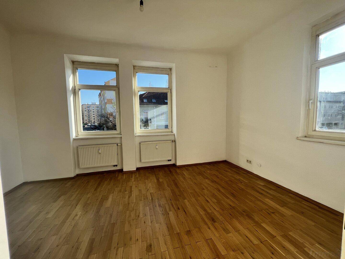 Geräumiges Wohnzimmer mit hellem Holzboden und großen Fenstern, die viel Licht hereinlassen.
