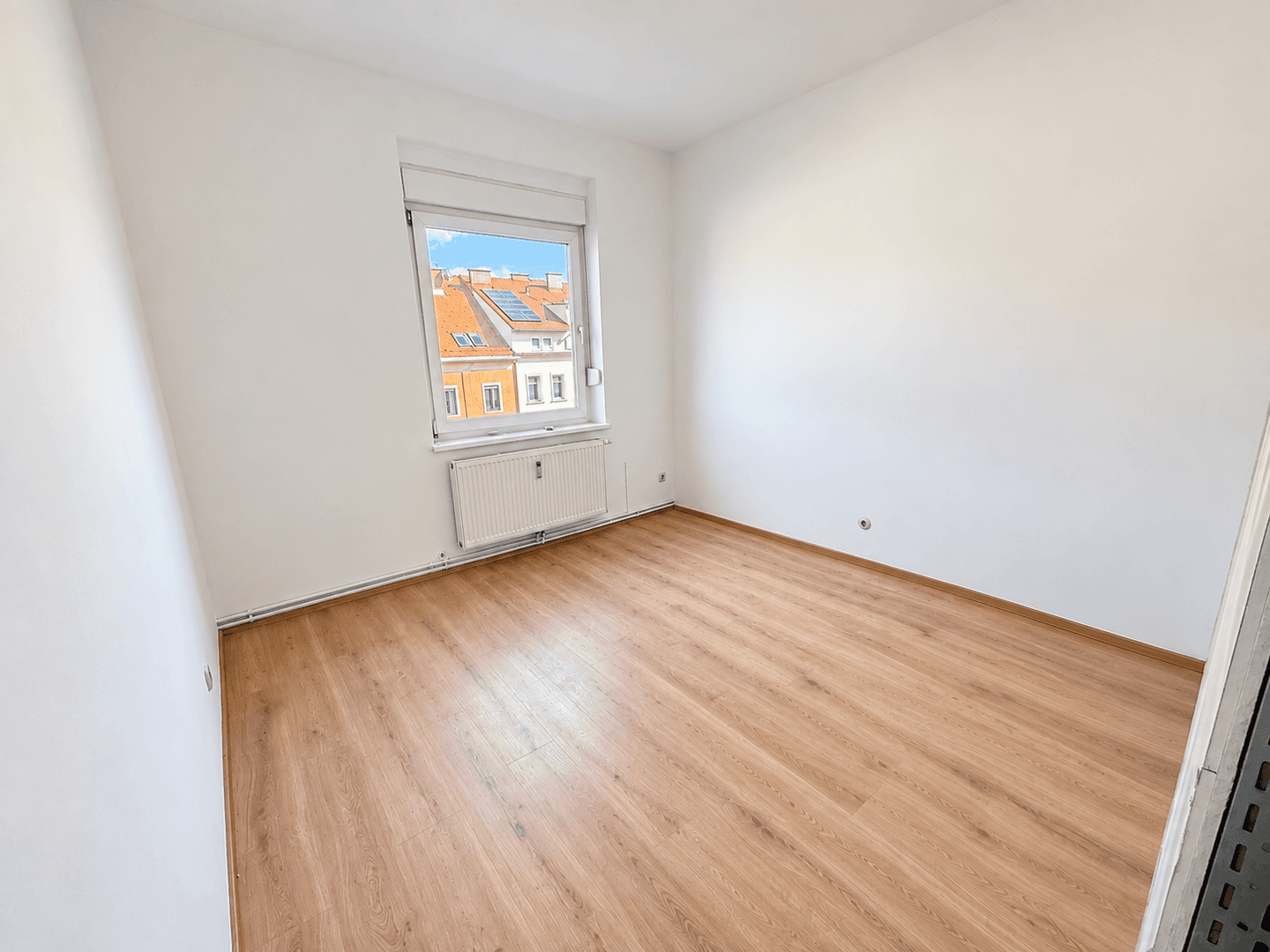Helles Zimmer mit Laminatboden, weißen Wänden und Blick auf die umliegenden Dächer.