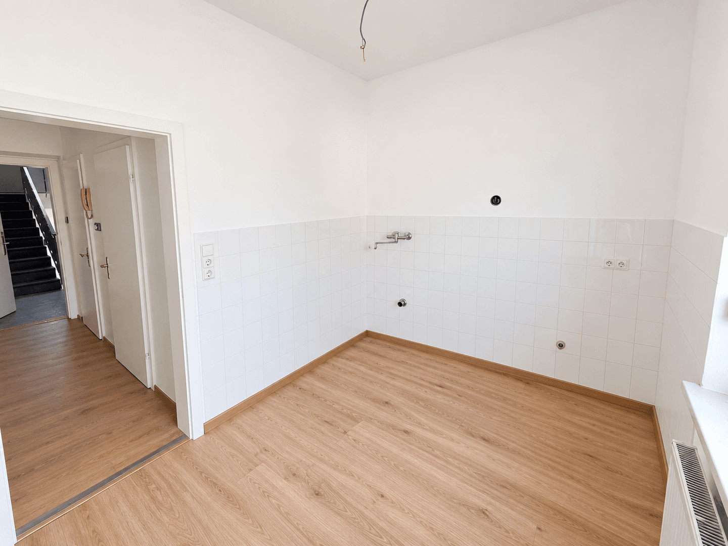 Leerer Küchenbereich mit gefliesten Wänden und Anschlüssen, offen zum Flur mit Treppenhaus.