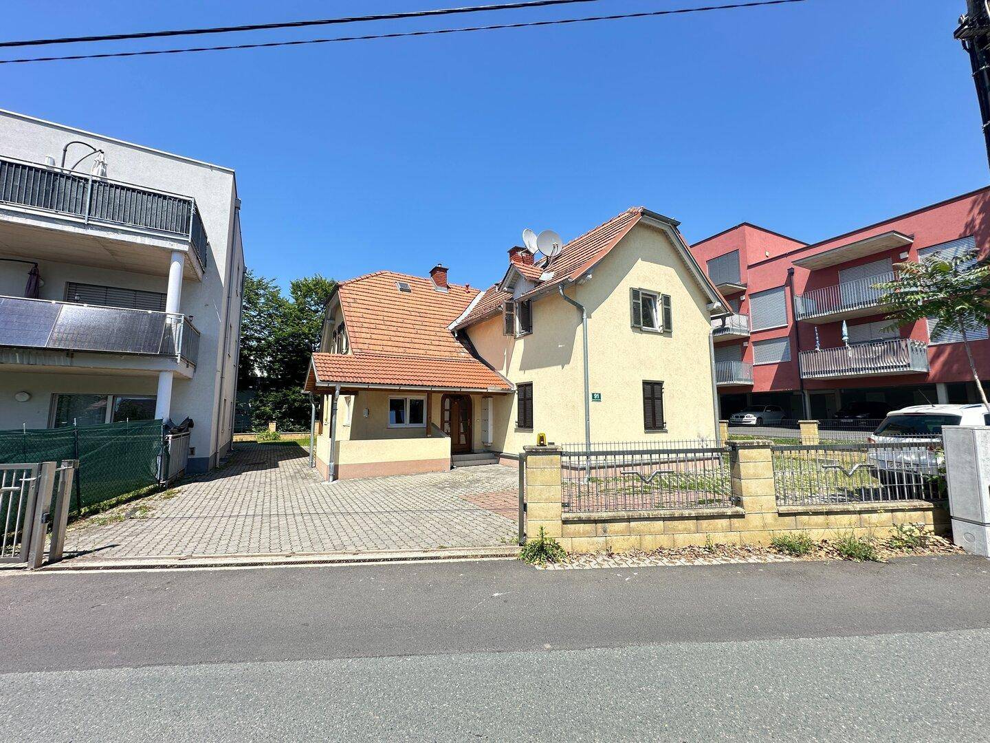 Traditionelles gelbes Haus mit Ziegeldach und überdachtem Eingang, umgeben von moderneren Gebäuden.