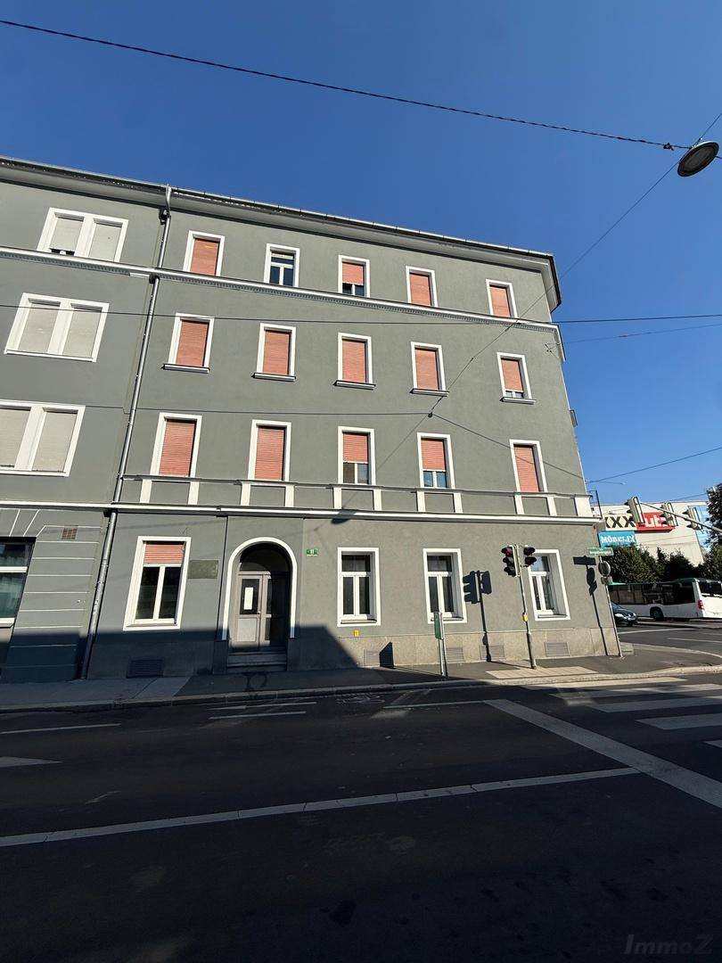 Graues Gebäude mit roten Fensterläden an einer Straßenecke unter blauem Himmel, mit moderner Architektur.