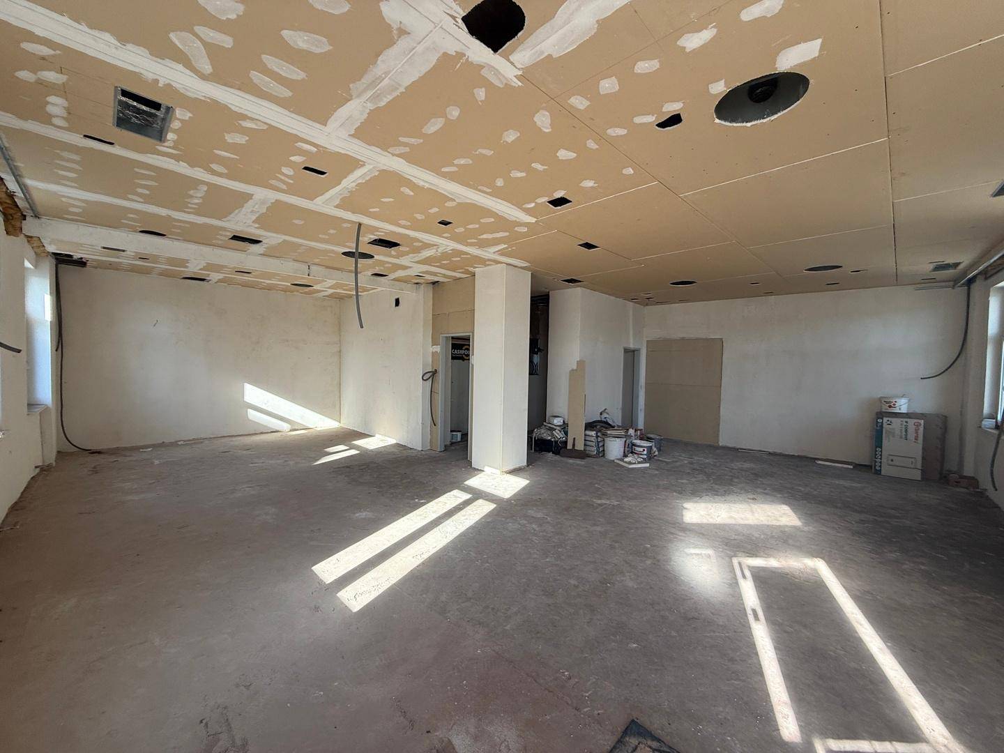 Weitläufiger Innenraum im Rohbau mit sichtbaren Deckenplatten und Betonboden, viel Tageslicht durch Fenster.