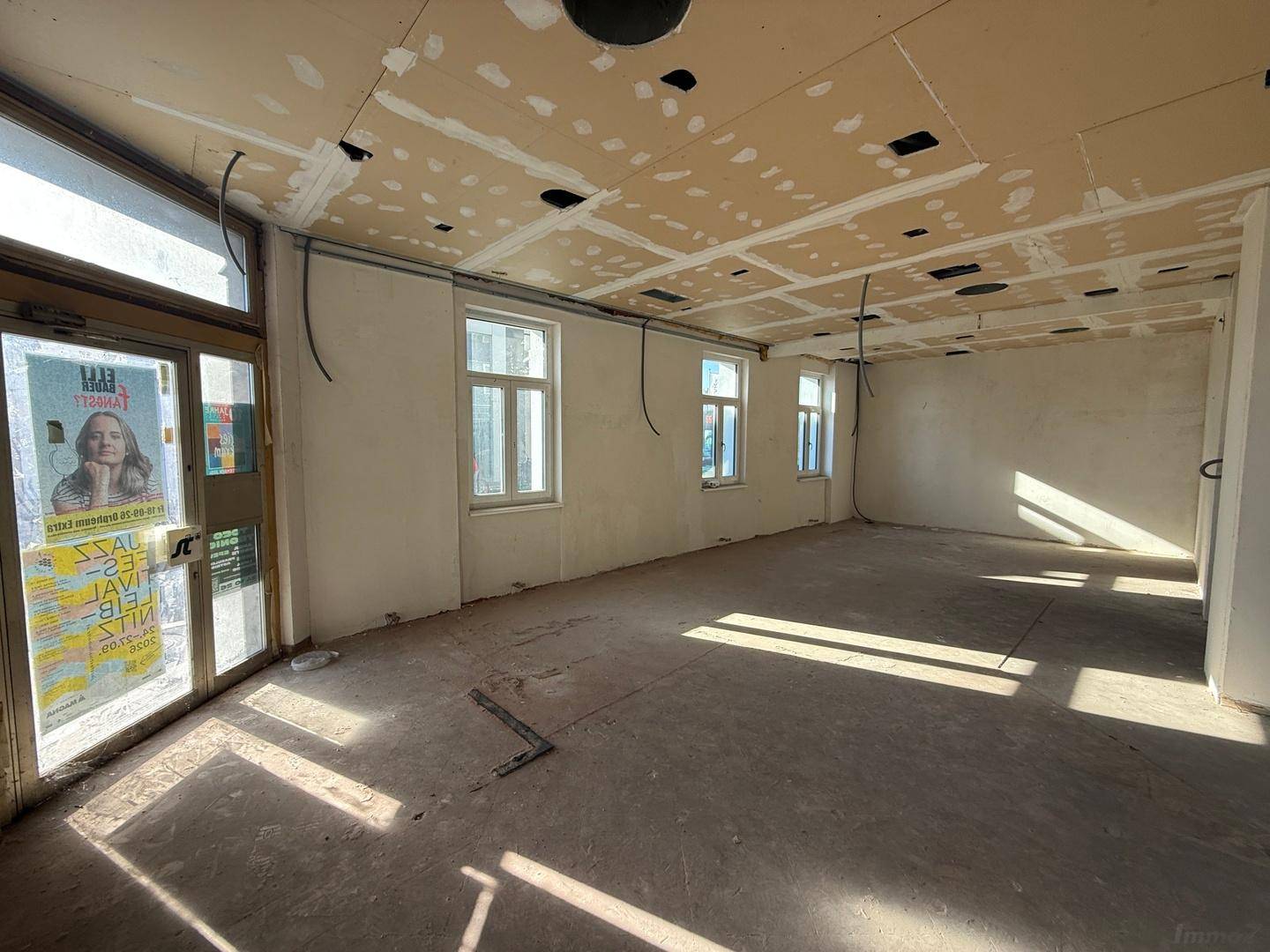 Unfertiger Innenraum mit Betonboden und großen Fenstern, die viel natürliches Licht hereinlassen, Blick zum Eingang.