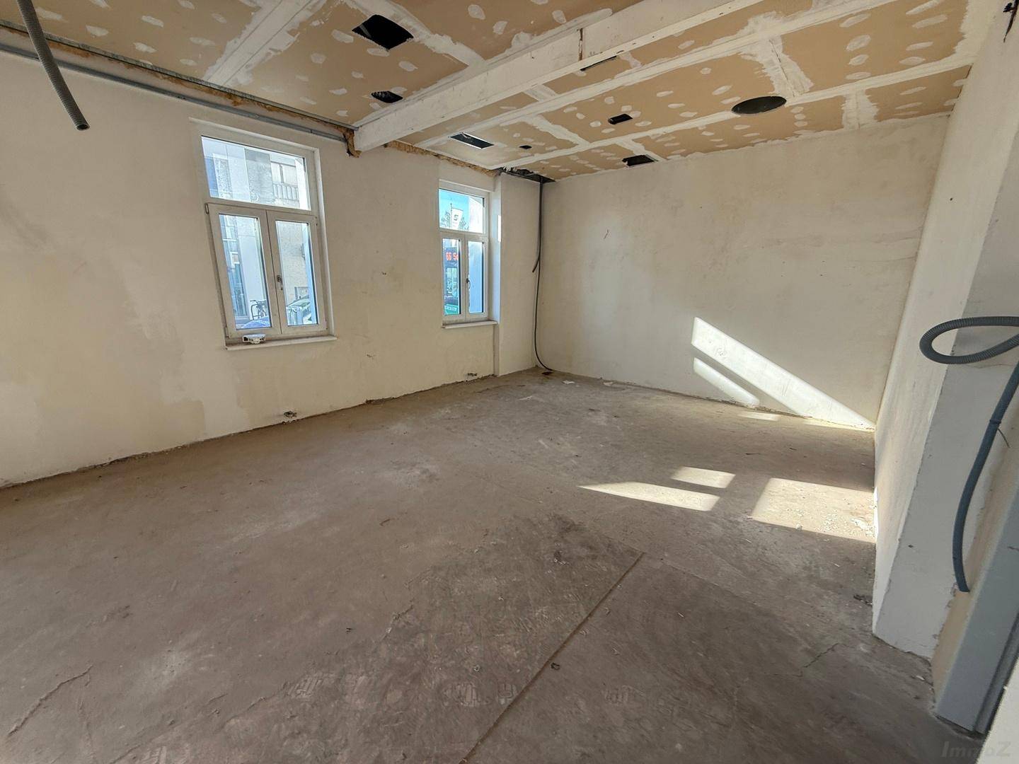 Heller Rohbau-Raum mit Betonboden und mehreren Fenstern, die den Raum mit natürlichem Licht durchfluten.