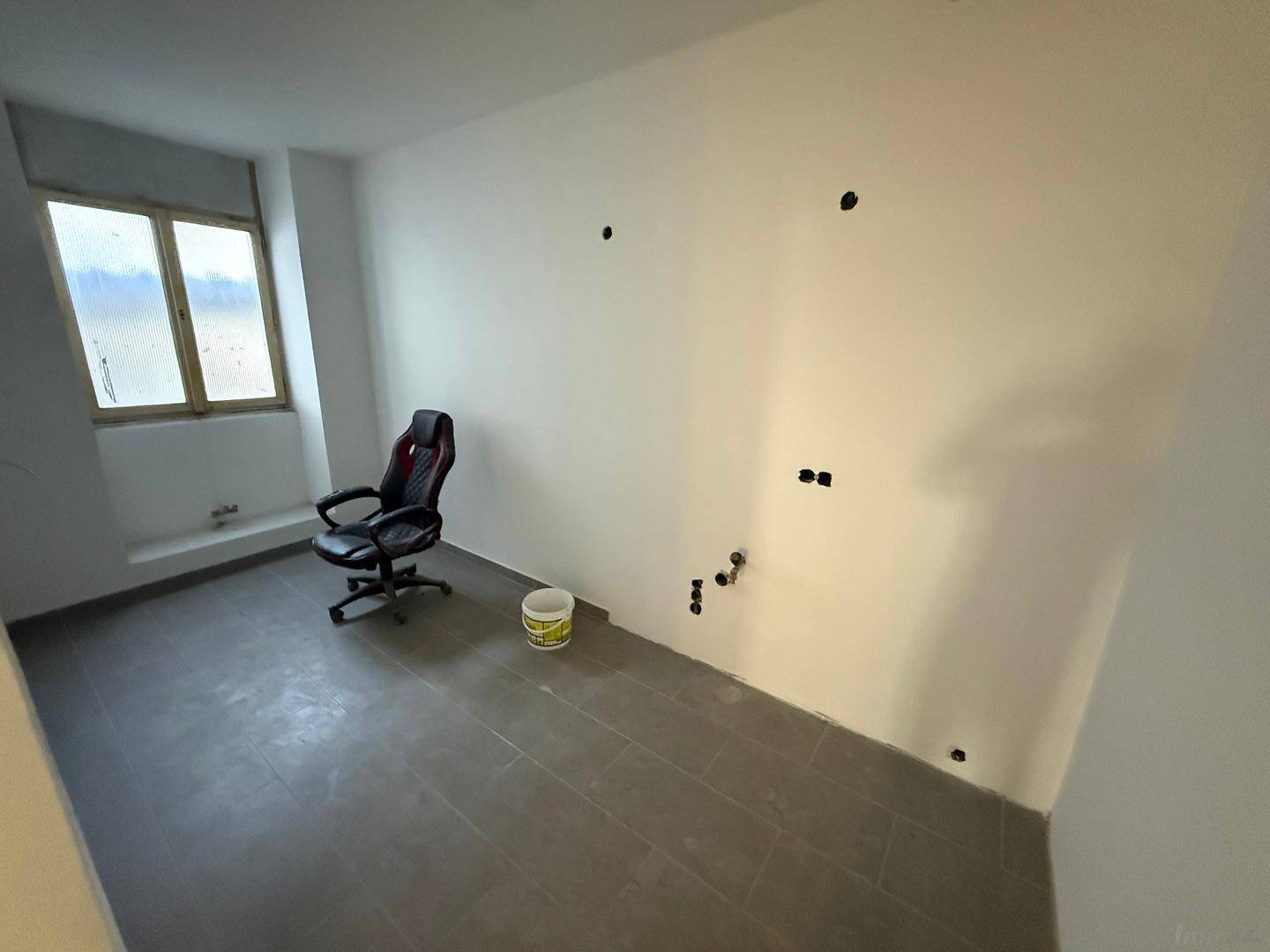 Helles Büro mit grauen Fliesen und weißen Wänden, einem Fenster und vorbereiteten Elektroinstallationen.