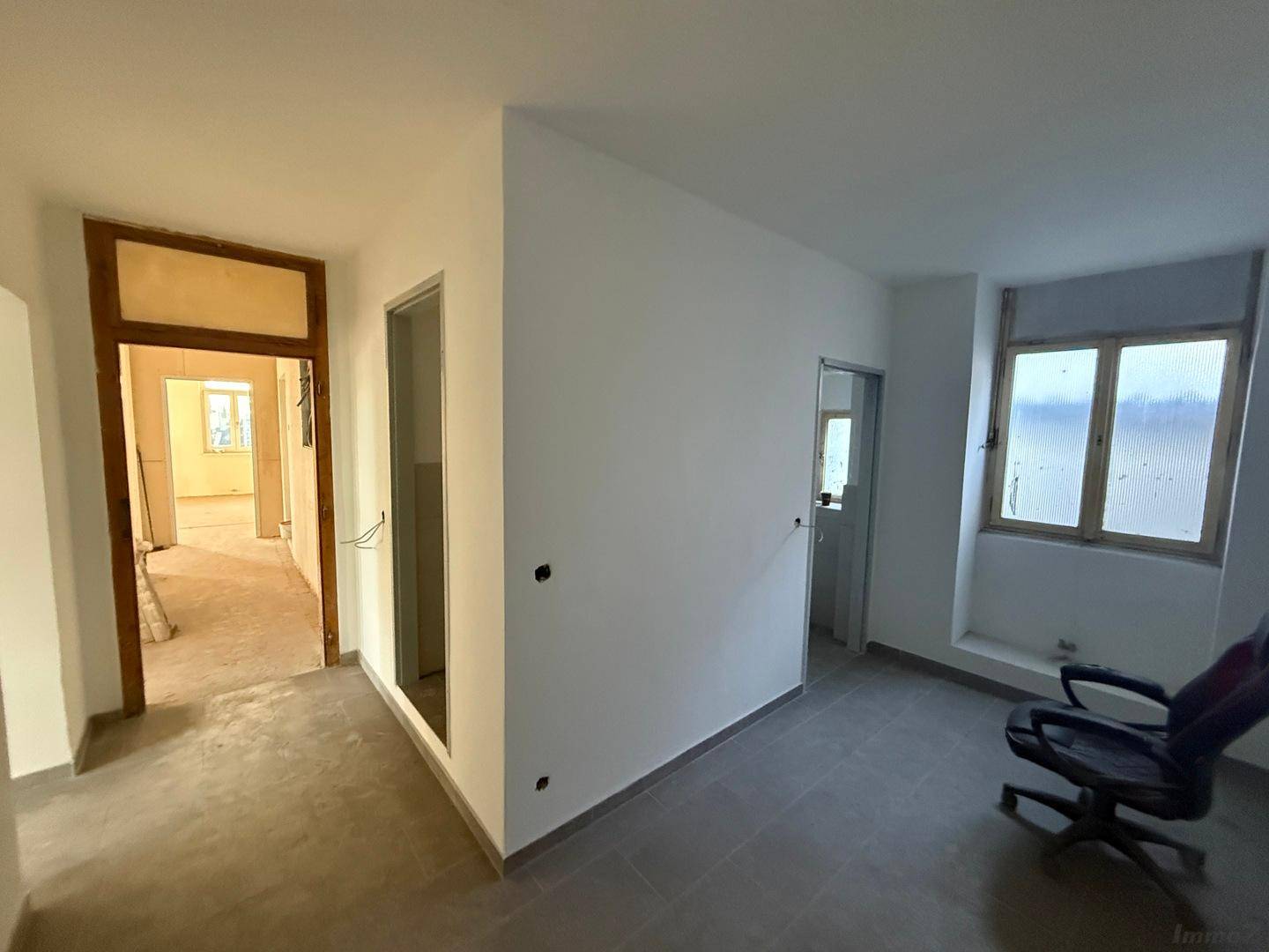 Büroähnlicher Raum mit grauen Fliesen, weißen Wänden und einem Fenster, das natürliches Licht spendet.