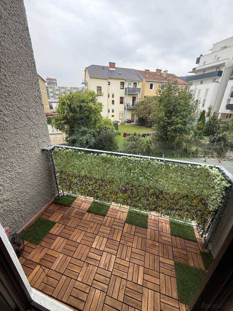 Balkon mit Holzbodenfliesen und künstlicher Begrünung, mit Blick auf umliegende Gebäude und Grünflächen.