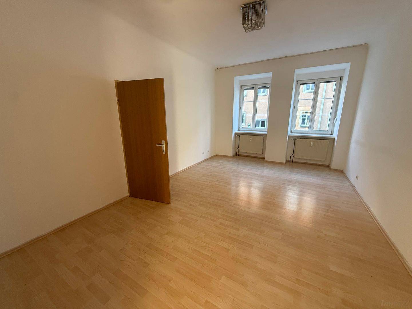Geräumiges, leeres Zimmer mit Laminatboden und zwei Fenstern, die den Raum erhellen.