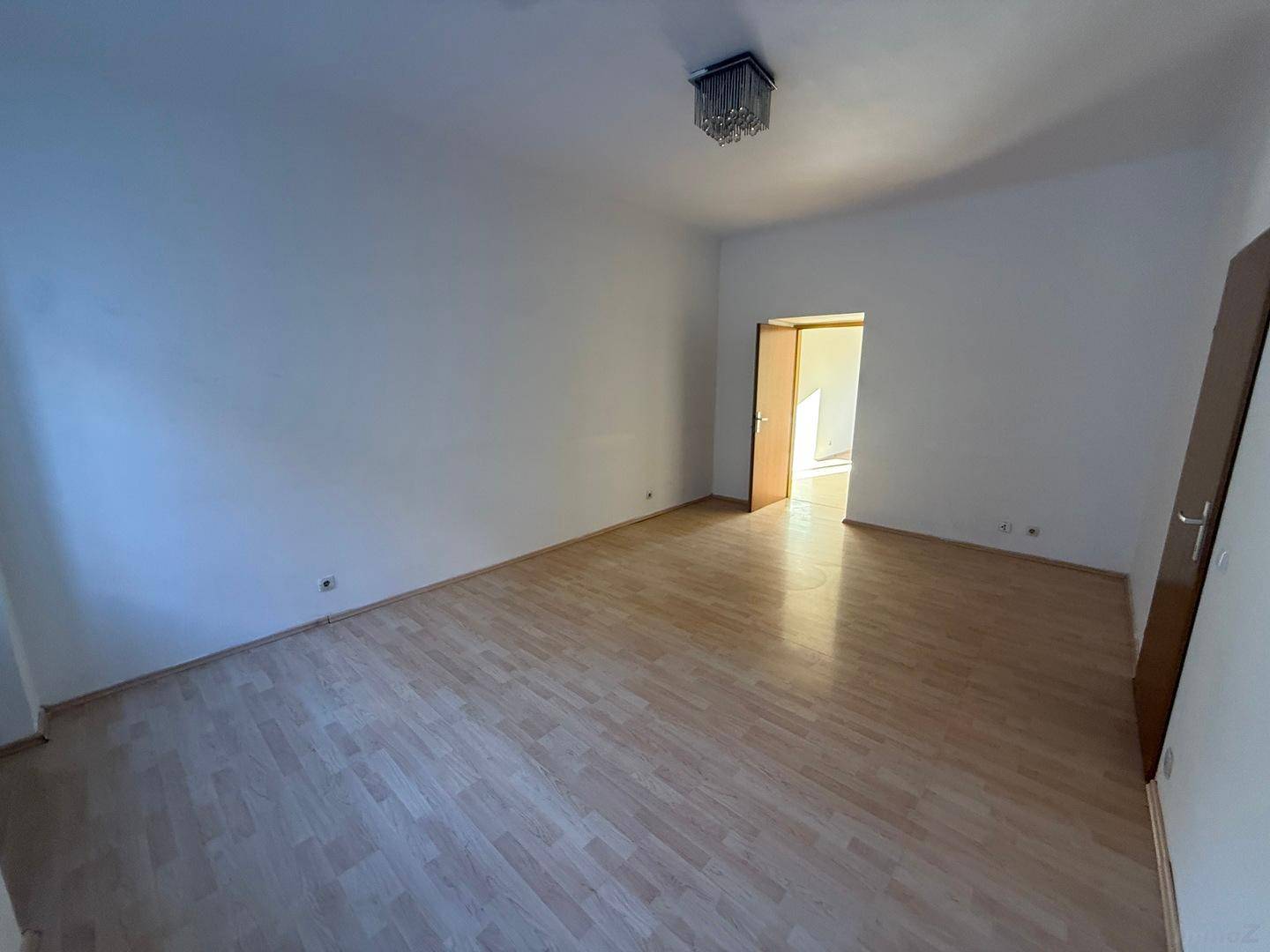 Heller, leerer Raum mit Laminatboden und einer Tür, die zu einem weiteren Zimmer führt.