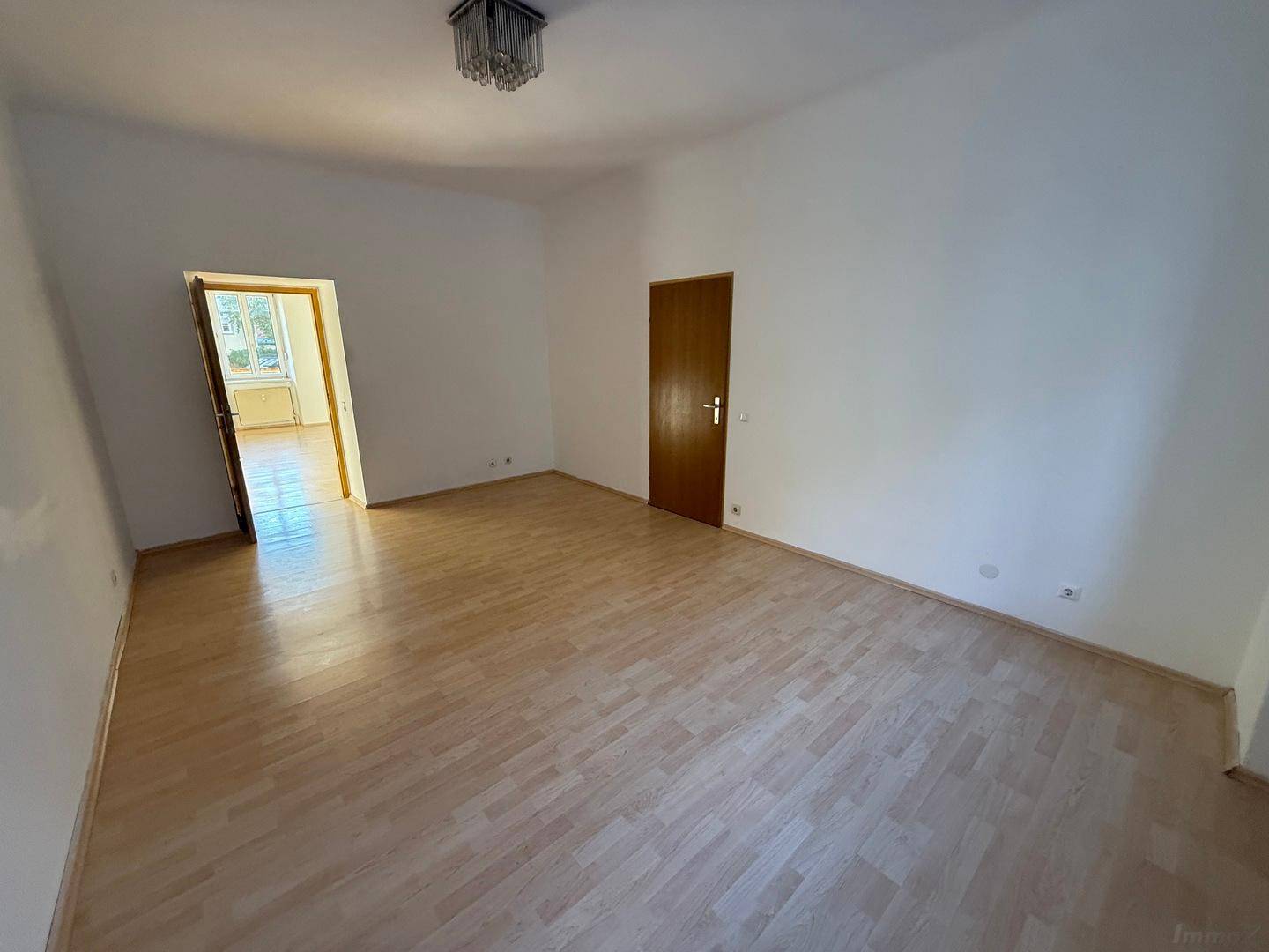 Leerer Wohnbereich mit Laminatboden und einer offenen Tür, die den Blick in einen weiteren Raum freigibt.