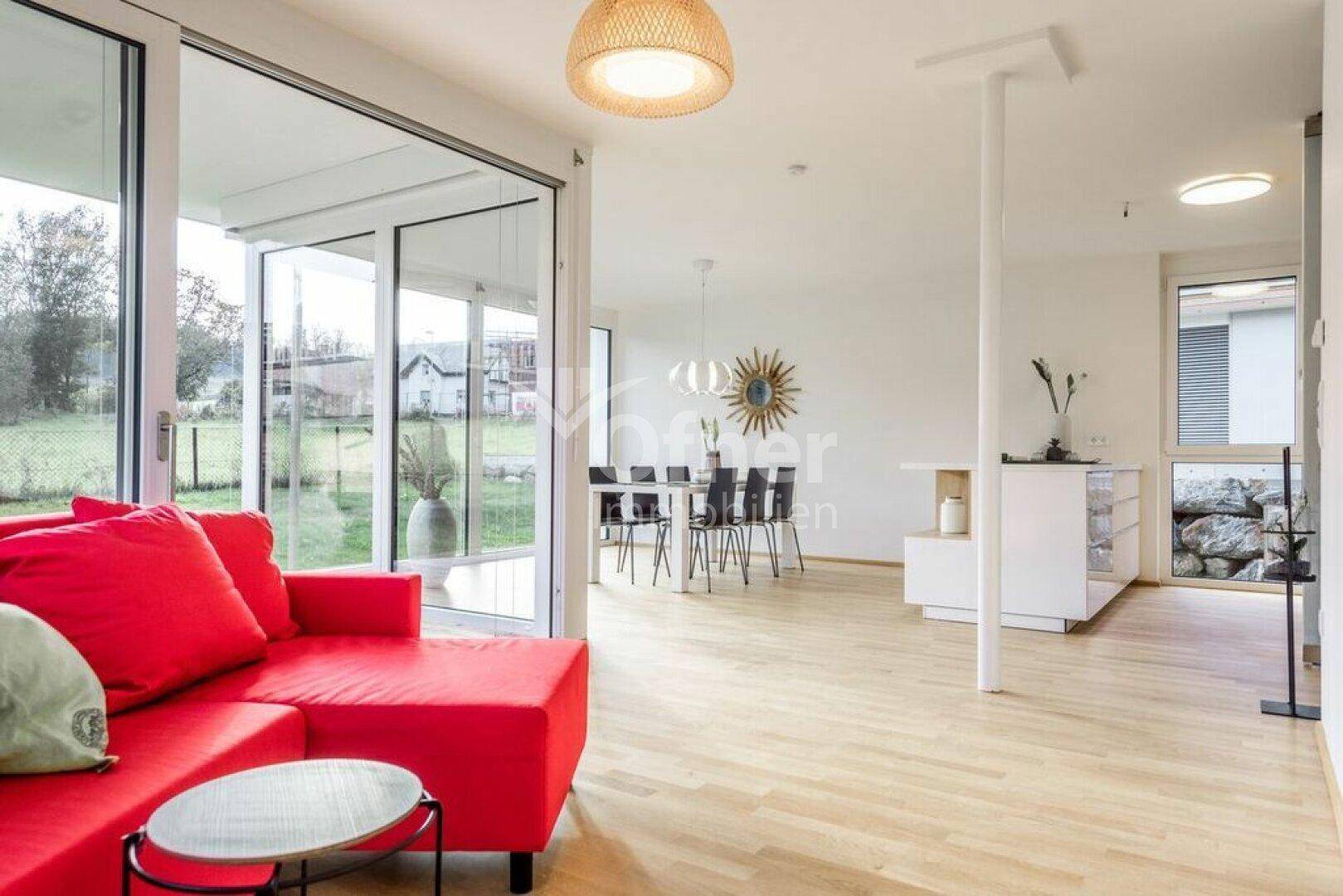 Heller offener Wohn-Essbereich mit rotem Sofa, Holzboden und großen Schiebetüren zur Terrasse.