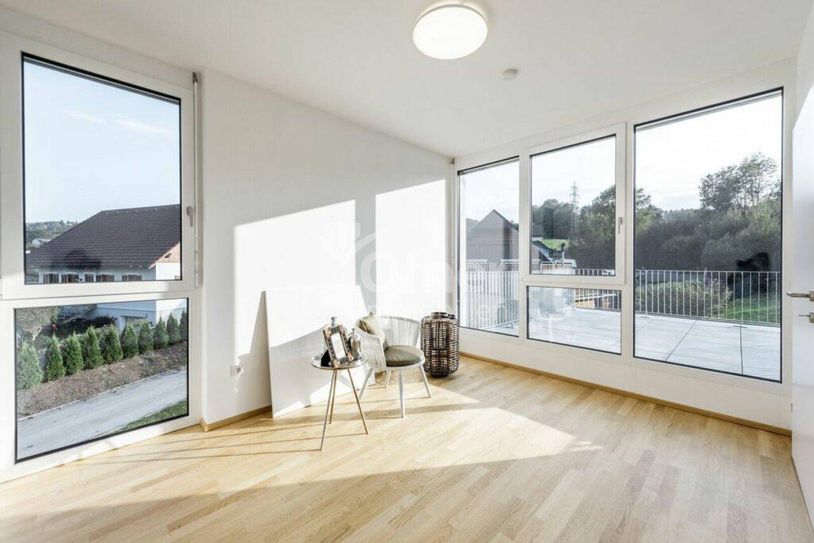 Lichtdurchfluteter Raum mit großen Fenstern, Holzboden und Zugang zu einer Dachterrasse.