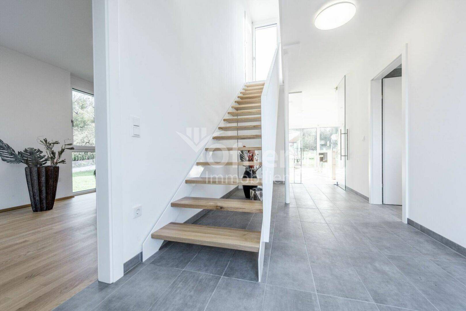 Heller Flur mit offener Holztreppe, grauen Bodenfliesen und Zugang zu weiteren Räumen.