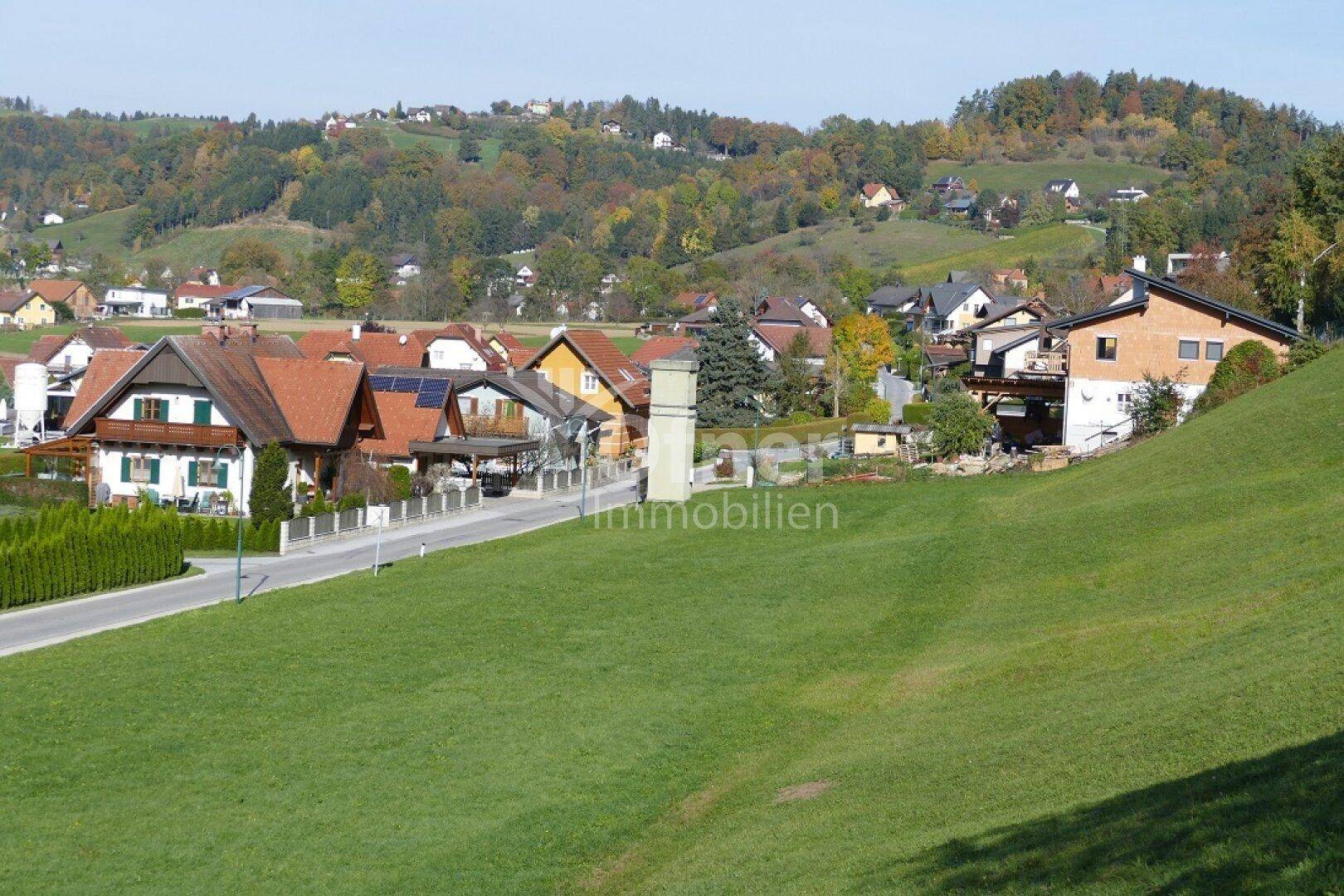 Weitläufige Ansicht einer Wohngegend mit Häusern, Gärten und bewaldeten Hügeln im Hintergrund.