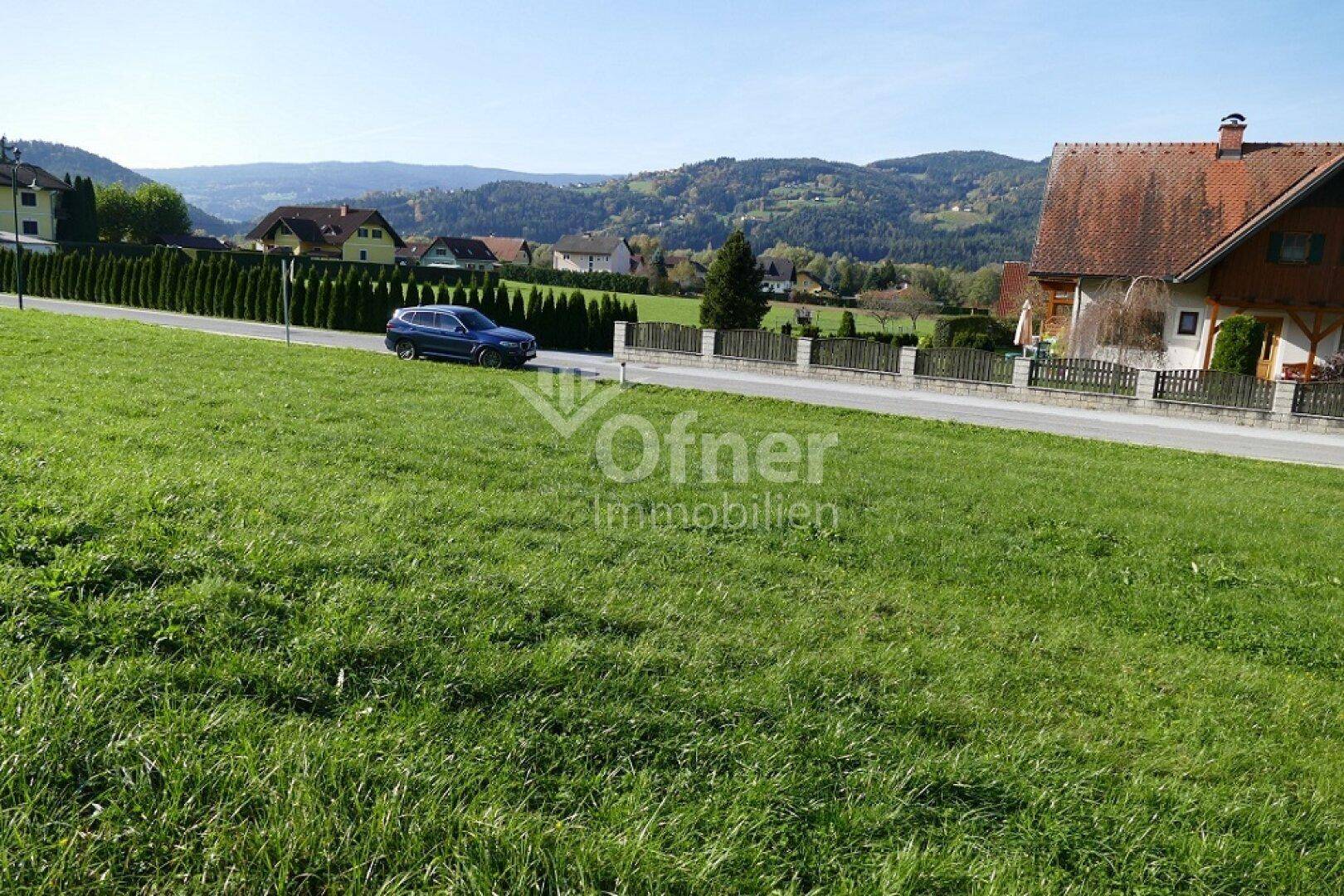 Grünes Grundstück mit einer Straße, Hecke und Häusern im Hintergrund, umgeben von Hügeln.
