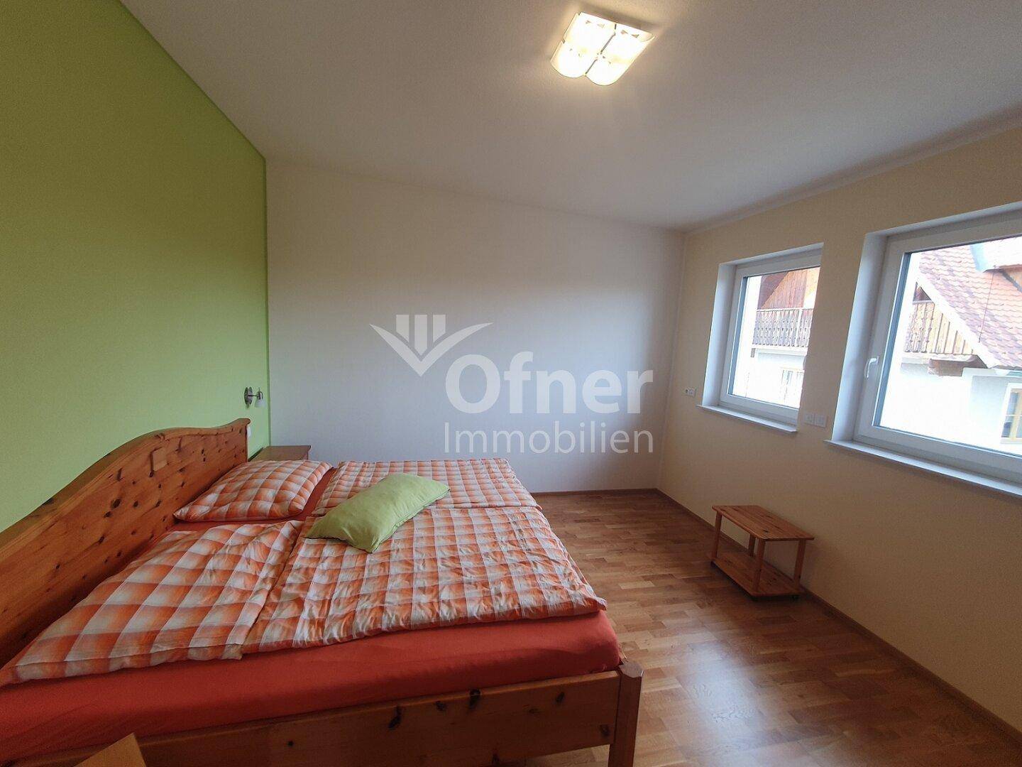 Schlafzimmer mit Holzbett, grüner Akzentwand und zwei Fenstern für viel Tageslicht.