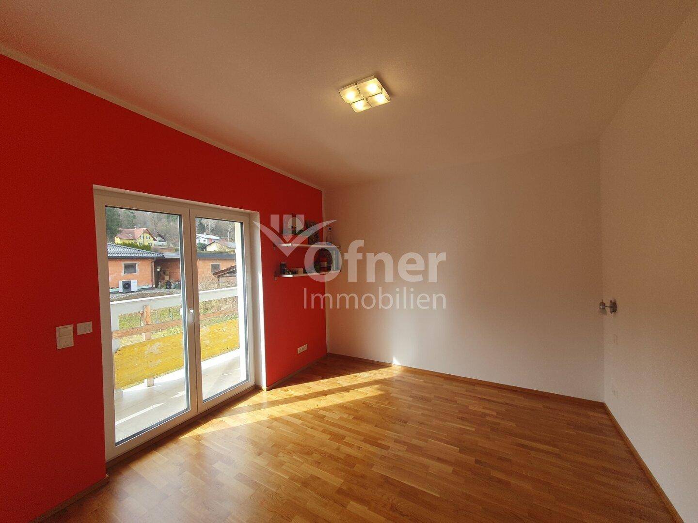 Leeres Zimmer mit roter Akzentwand, Holzboden und großem Fenster mit Zugang nach draußen.