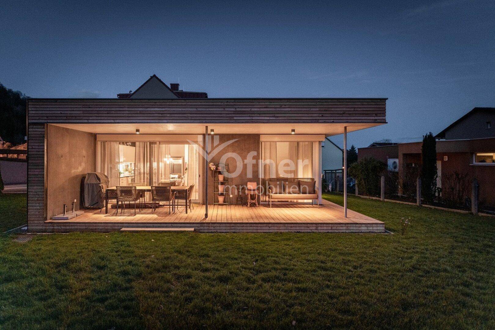 Beleuchtete Holzterrasse mit Außenmöbeln und Grillbereich, eingebettet in einen grünen Garten.