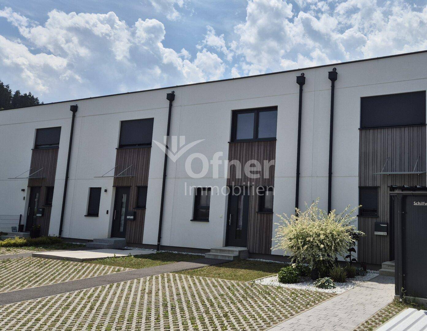 Reihe moderner Häuser mit weißer Fassade, dunklen Fenstern und Holzverkleidung unter bewölktem Himmel.