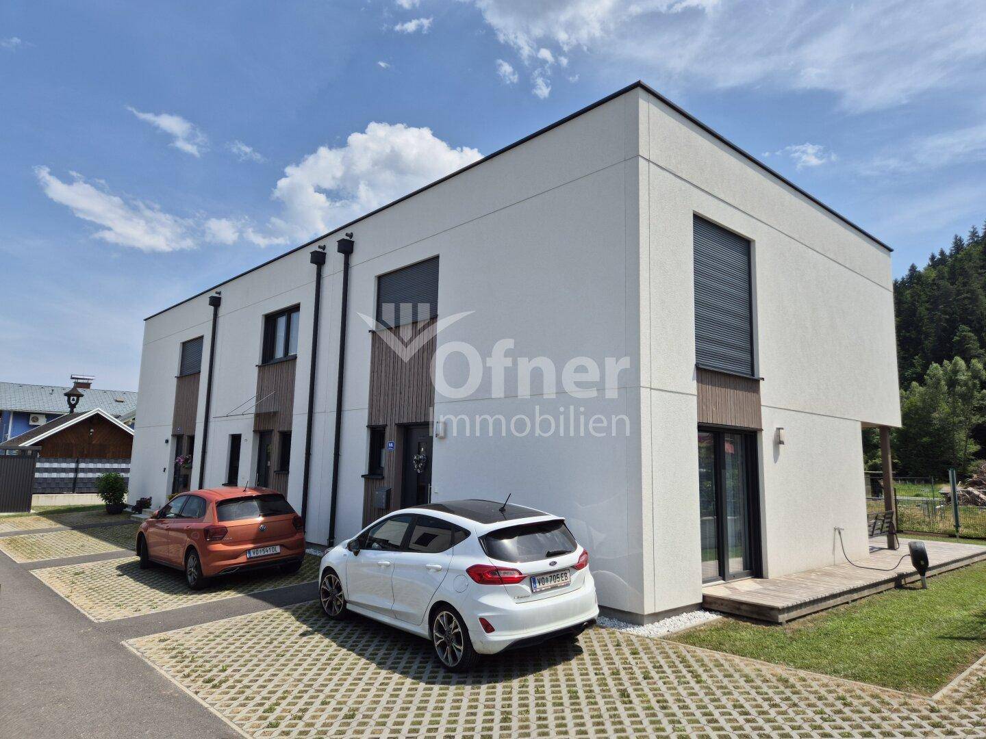 Modernes Reihenhaus mit weißer Fassade, Holzdetails und gepflasterten Parkplätzen davor.