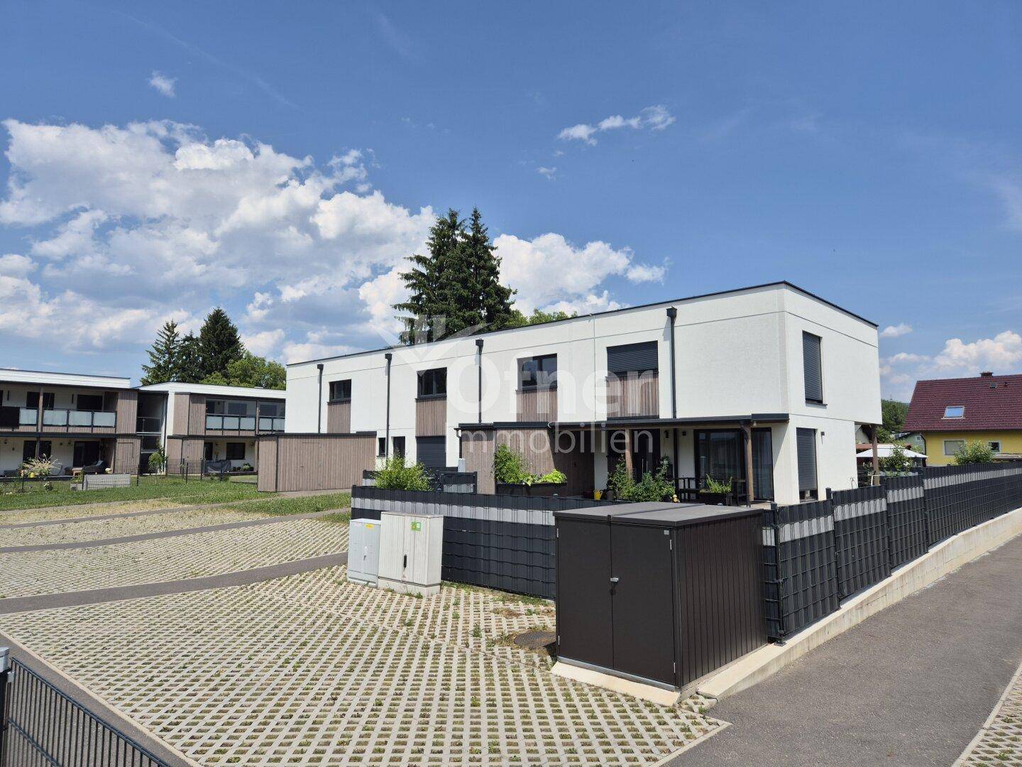 Moderne Reihenhäuser mit Parkplätzen und einem kleinen Gartenbereich unter blauem Himmel.