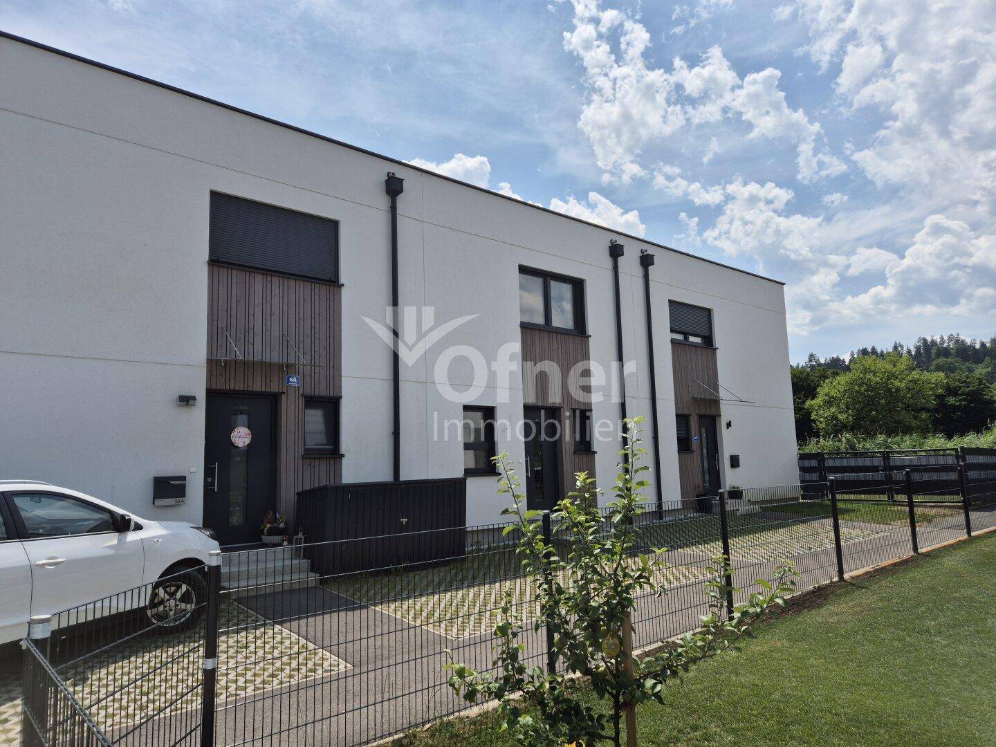 Vorderansicht eines modernen Reihenhauses mit weißer Fassade, dunklen Fenstern und gepflastertem Zugang.
