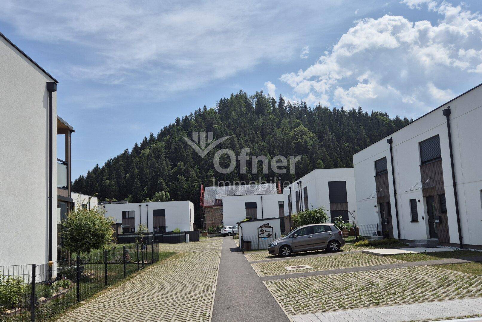 Moderne Wohnanlage mit Reihenhäusern und Blick auf bewaldete Hügel unter blauem Himmel.