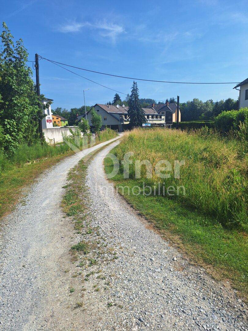 Schotterweg führt durch eine grüne Landschaft mit Häusern im Hintergrund unter blauem Himmel.
