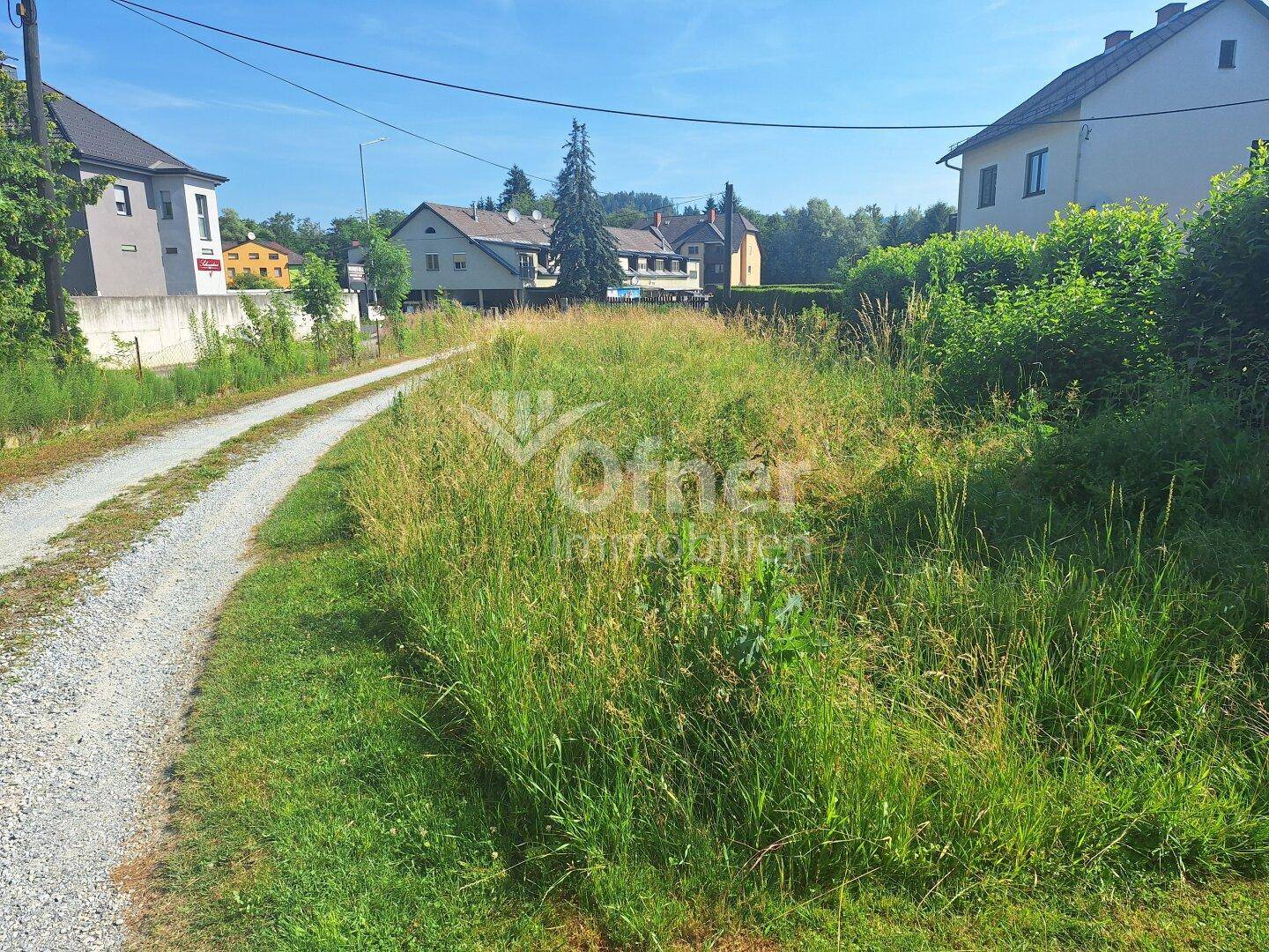 Breiter Schotterweg mit üppigem Gras am Rand, umgeben von ländlicher Bebauung und Natur.