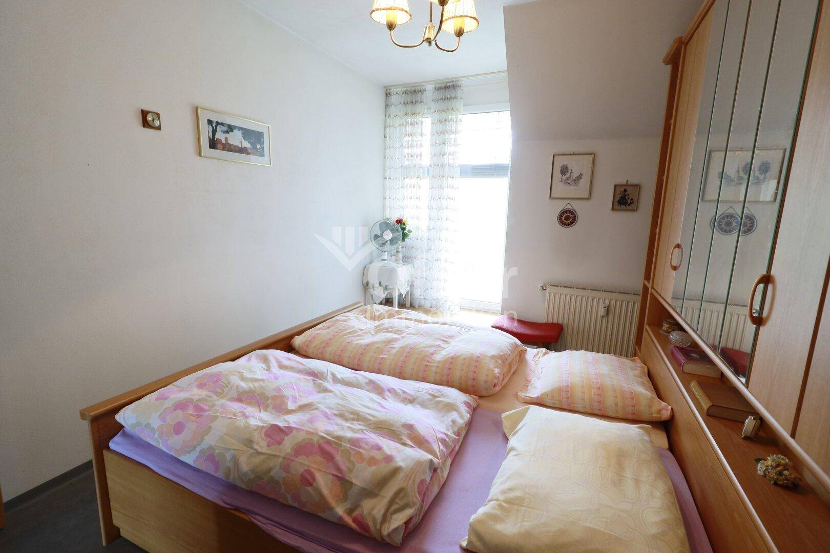 Schlafzimmer mit Doppelbett, Kleiderschrank und Fenster, das viel Tageslicht hereinlässt.