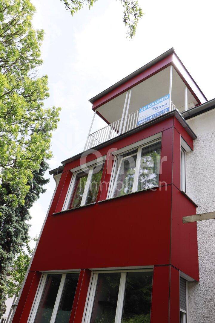 Seitenansicht eines Gebäudes mit roter Fassadenverkleidung und einem Balkon mit "Zu Kaufen"-Schild.