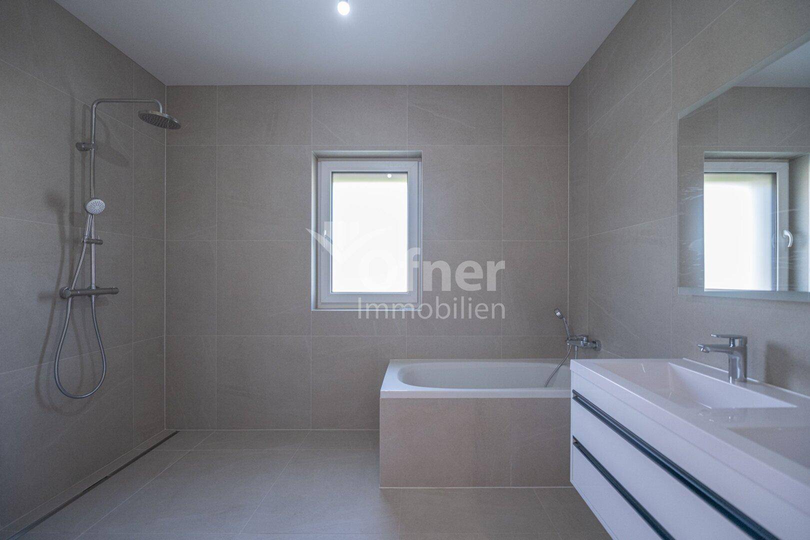 Modernes Badezimmer mit Badewanne, Dusche, Waschtisch und Fenster in hellgrauen Fliesen.