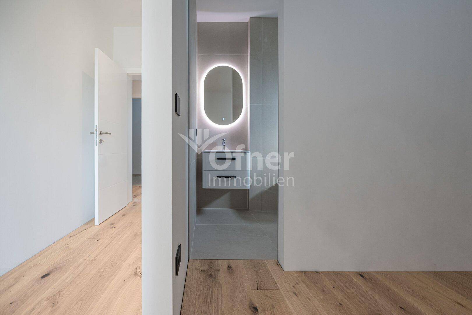 Flur mit Blick in ein modernes Badezimmer, das einen beleuchteten Spiegel und Waschtisch zeigt.