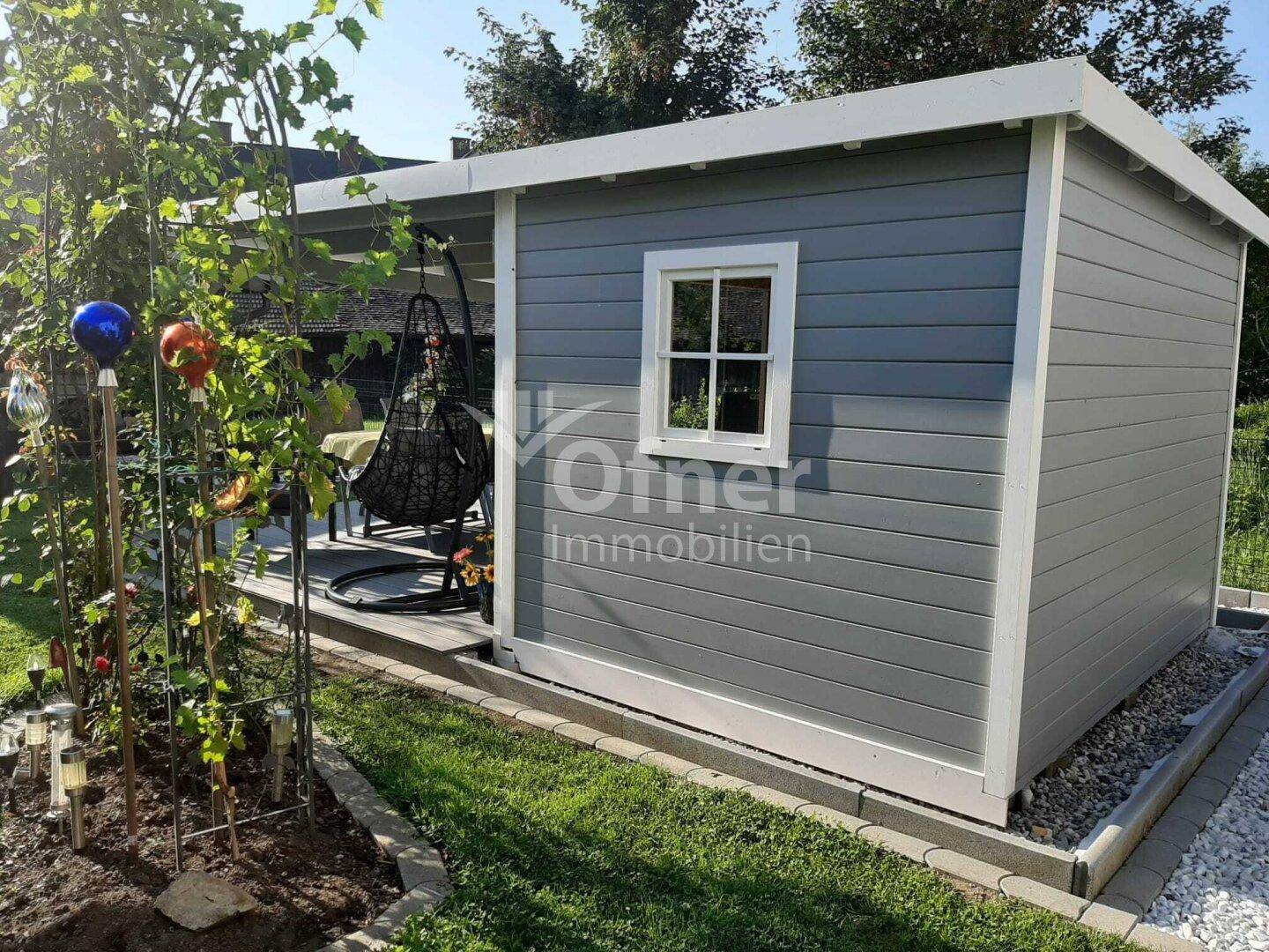 Modernes Gartenhaus in Grau mit kleiner Terrasse und Schaukelstuhl im Garten.