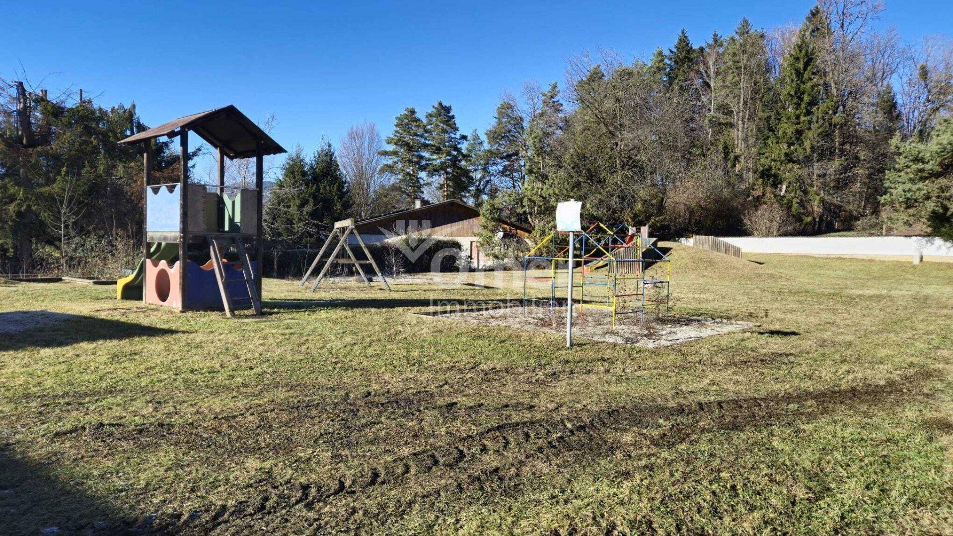 Sonniger Spielplatz mit Rutsche, Klettergerüst und Schaukeln auf einer weitläufigen Wiese.