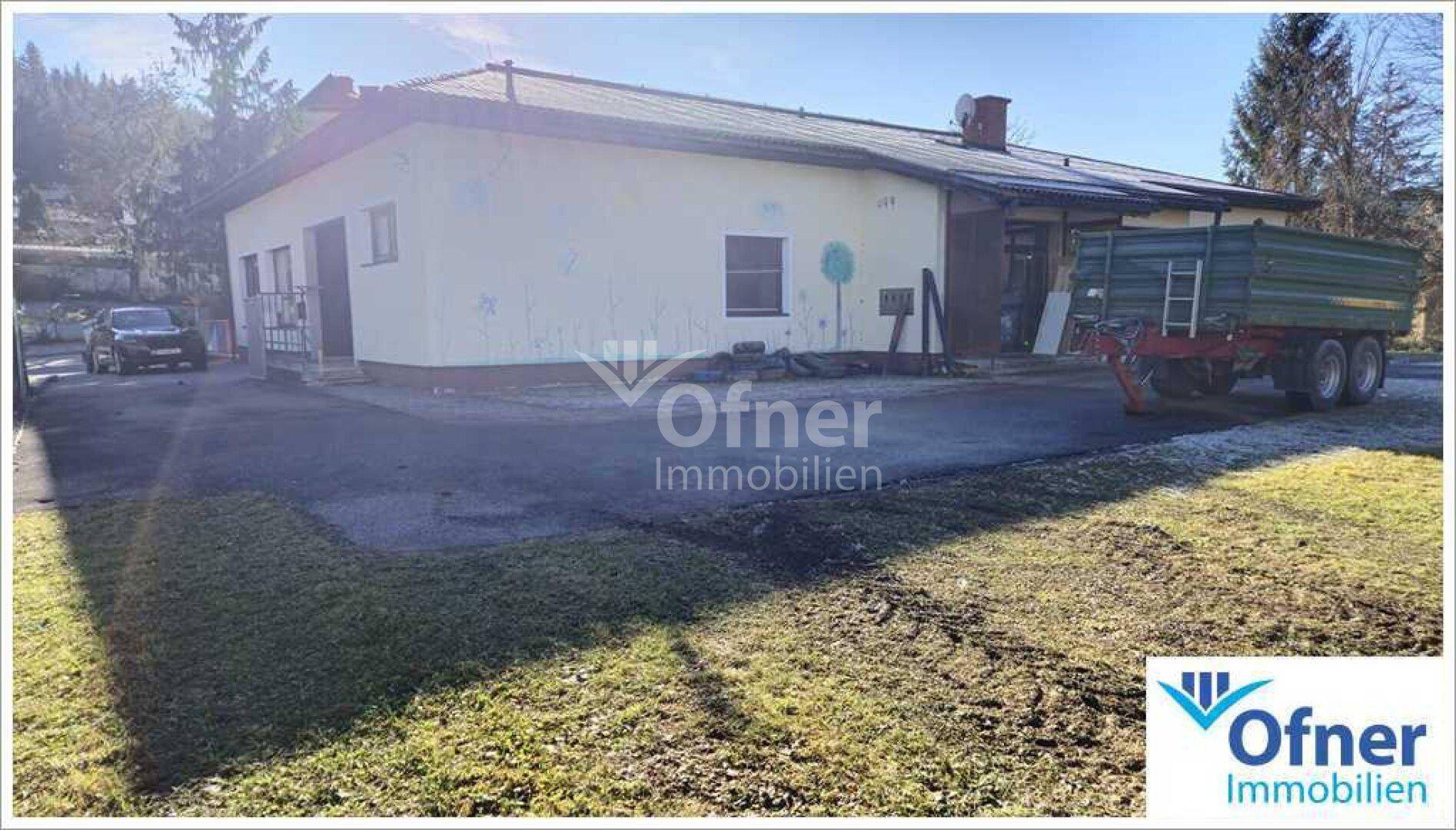 Gebäudeansicht mit gepflastertem Vorplatz und einem Anhänger, umgeben von Grünflächen.