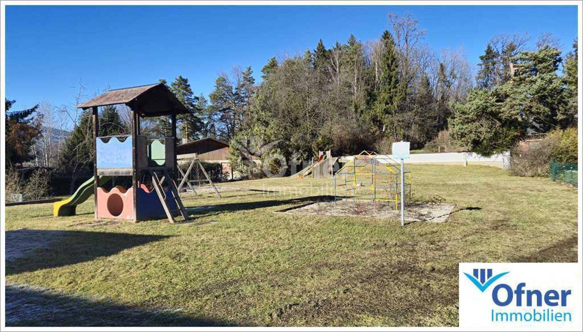 Weitläufiger Spielplatz mit verschiedenen Geräten und umgebenden Bäumen an einem sonnigen Tag.