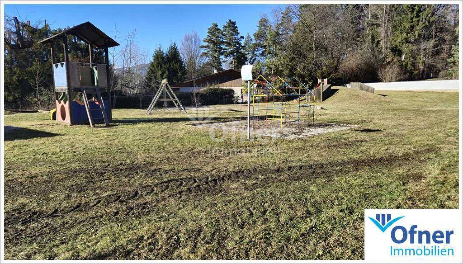 Großer Spielplatz mit Klettergerüst und Rutsche, umgeben von Bäumen und weitem Rasen.