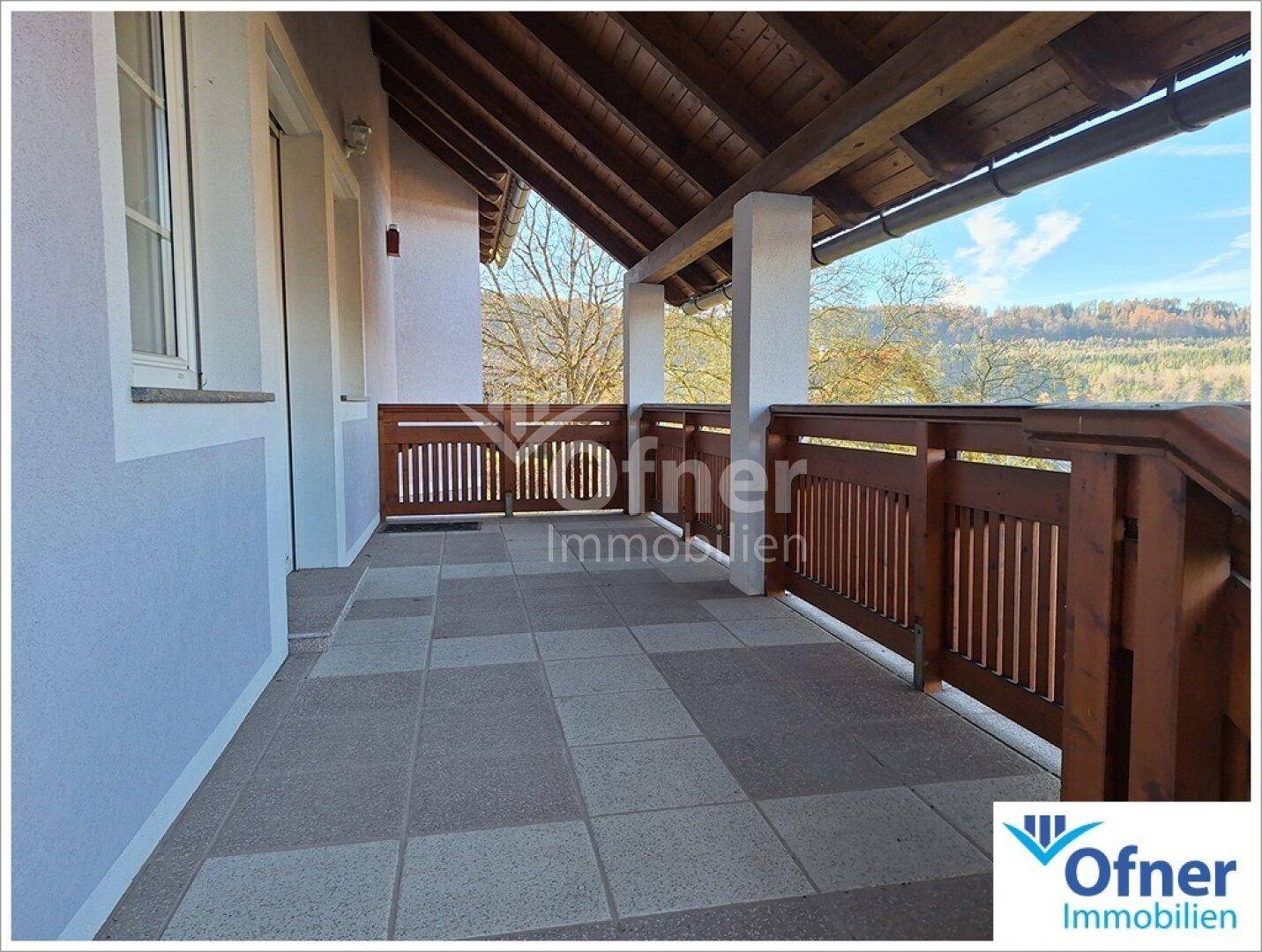 Überdachter Balkon mit Fliesenboden und Holzgeländer, bietet einen weiten Blick ins Grüne.